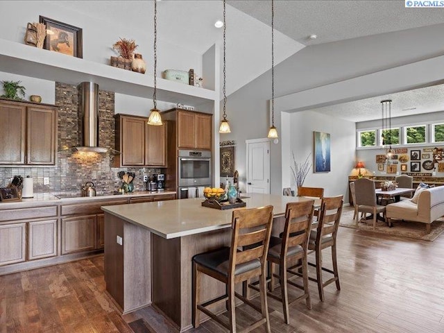 kitchen with brown cabinetry, double oven, wall chimney range hood, a center island, and light countertops