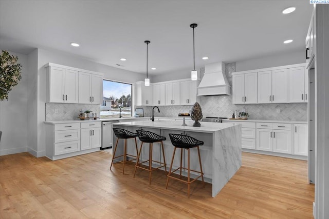 kitchen with premium range hood, white cabinetry, a kitchen island with sink, stainless steel dishwasher, and light hardwood / wood-style flooring