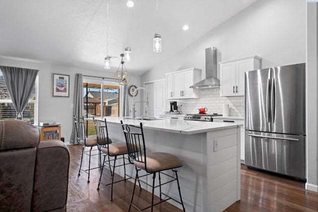 kitchen with stainless steel refrigerator, decorative light fixtures, white cabinetry, a kitchen island with sink, and wall chimney range hood