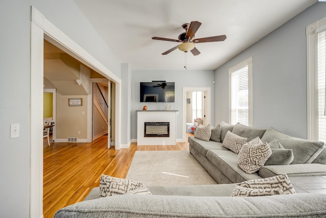 living area with visible vents, a glass covered fireplace, ceiling fan, wood finished floors, and baseboards