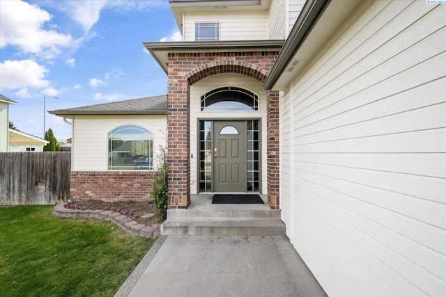 view of exterior entry with brick siding, fence, and a shingled roof