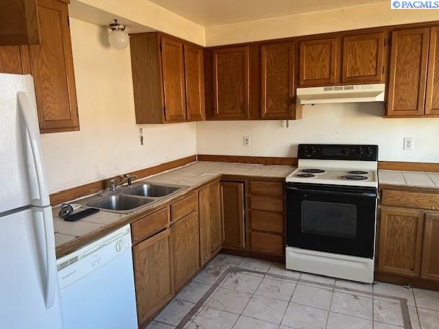 kitchen with under cabinet range hood, white appliances, a sink, tile counters, and brown cabinets