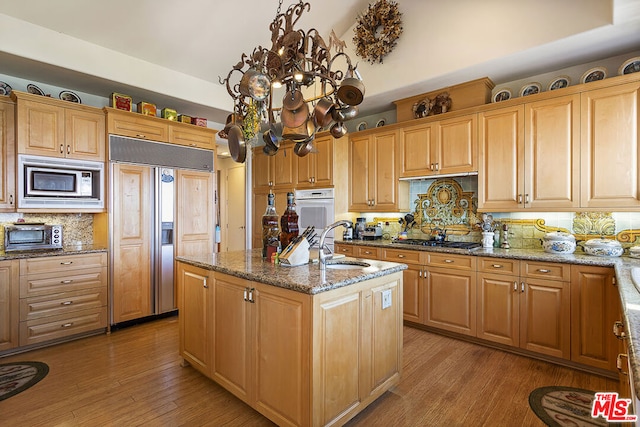 kitchen with light hardwood / wood-style flooring, a kitchen island with sink, built in appliances, and stone counters