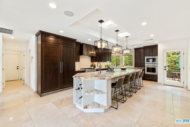 kitchen featuring dark brown cabinetry, light stone countertops, decorative light fixtures, a kitchen island with sink, and custom range hood