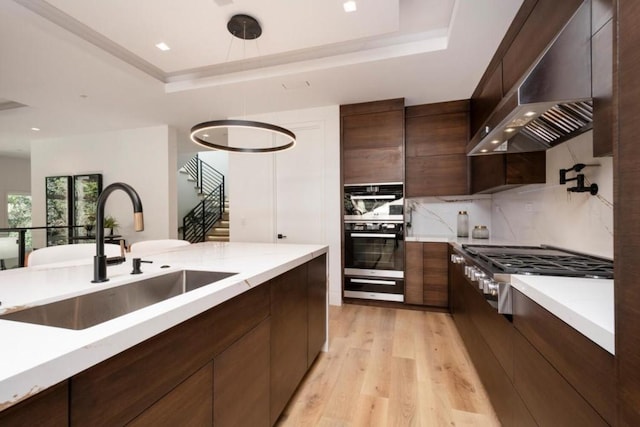 kitchen featuring a tray ceiling, double oven, sink, light wood-type flooring, and wall chimney range hood