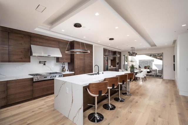 kitchen with stainless steel gas cooktop, a kitchen island with sink, light wood-type flooring, wall chimney range hood, and pendant lighting