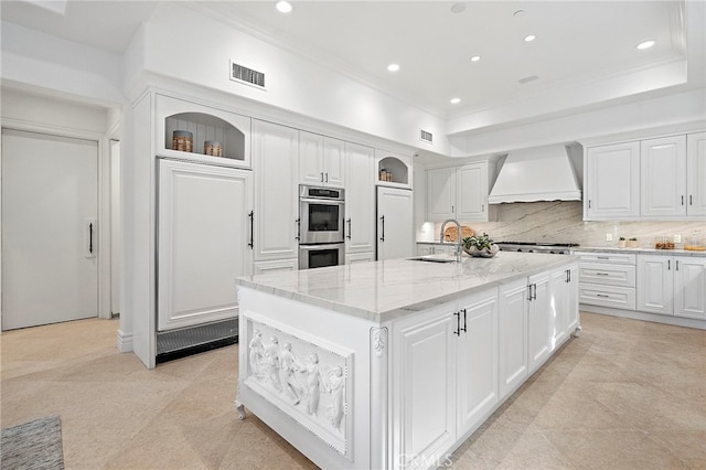 kitchen with premium range hood, light stone countertops, a center island with sink, white cabinets, and backsplash