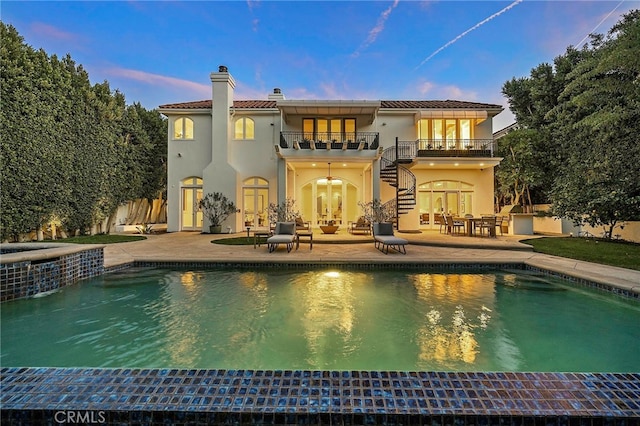 back house at dusk with a balcony, a patio, and a swimming pool with hot tub