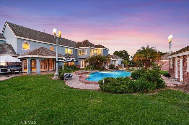 pool at dusk featuring a lawn, an in ground hot tub, and a patio