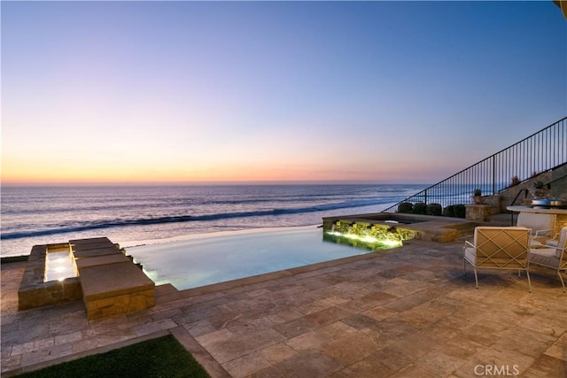 pool at dusk featuring a water view, a patio, and a beach view