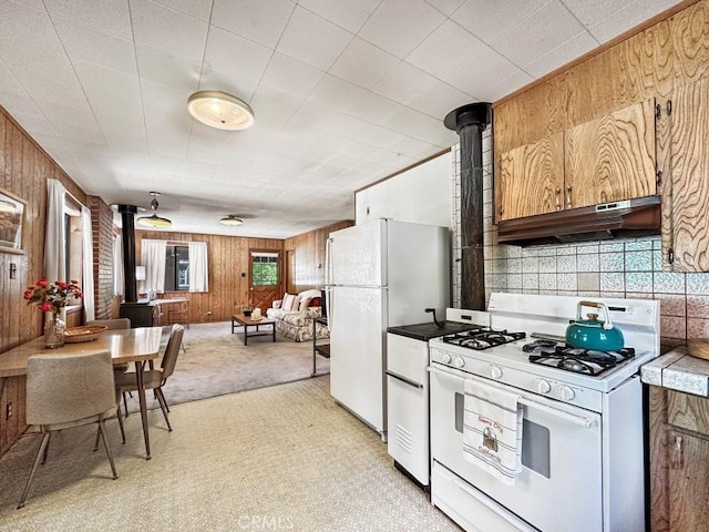 kitchen with a wood stove, white appliances, exhaust hood, light carpet, and wooden walls