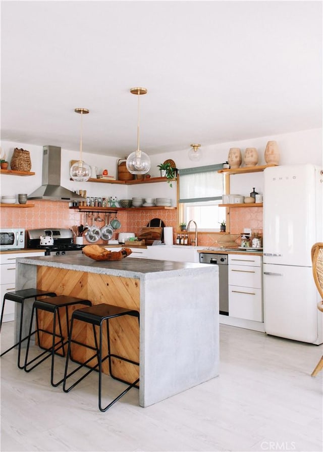 kitchen featuring appliances with stainless steel finishes, wall chimney range hood, pendant lighting, white cabinetry, and a breakfast bar area