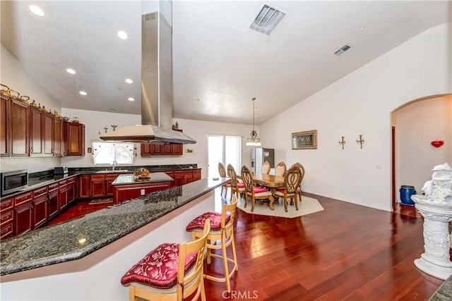 kitchen featuring dark hardwood / wood-style floors, pendant lighting, a kitchen breakfast bar, dark stone countertops, and island range hood