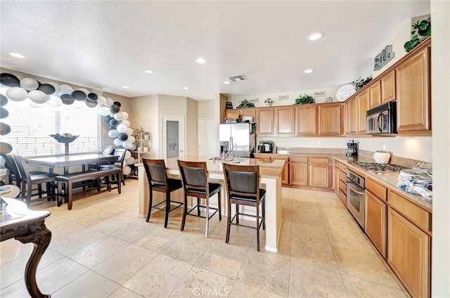 kitchen with a kitchen island with sink, light tile floors, appliances with stainless steel finishes, a breakfast bar area, and light stone countertops