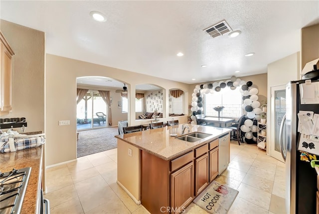 kitchen with stainless steel fridge, a kitchen island with sink, sink, light tile flooring, and light stone countertops