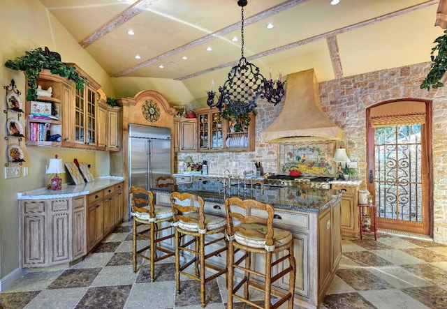 kitchen featuring wall chimney range hood, stainless steel built in refrigerator, dark stone counters, a breakfast bar area, and a center island with sink