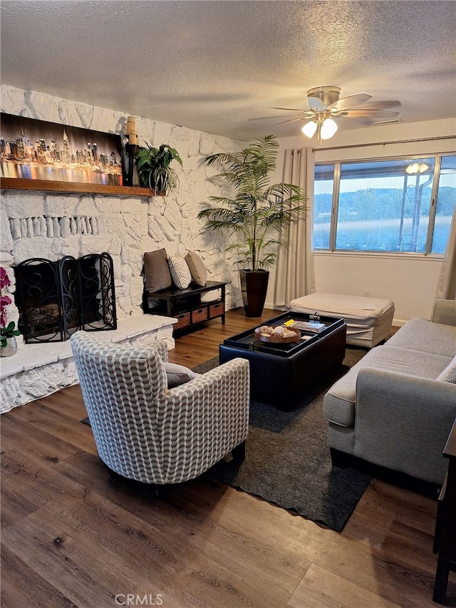 living room featuring hardwood / wood-style flooring, ceiling fan, a stone fireplace, and a textured ceiling