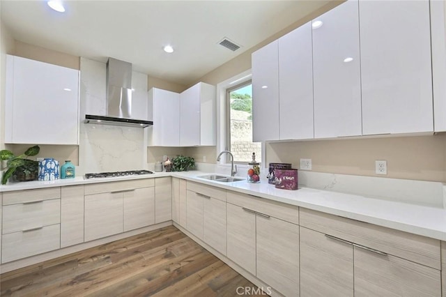 kitchen featuring white cabinetry, sink, wall chimney range hood, stainless steel gas stovetop, and hardwood / wood-style flooring