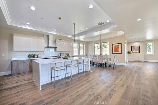 kitchen with white cabinets, a center island with sink, wall chimney range hood, a tray ceiling, and light hardwood / wood-style floors