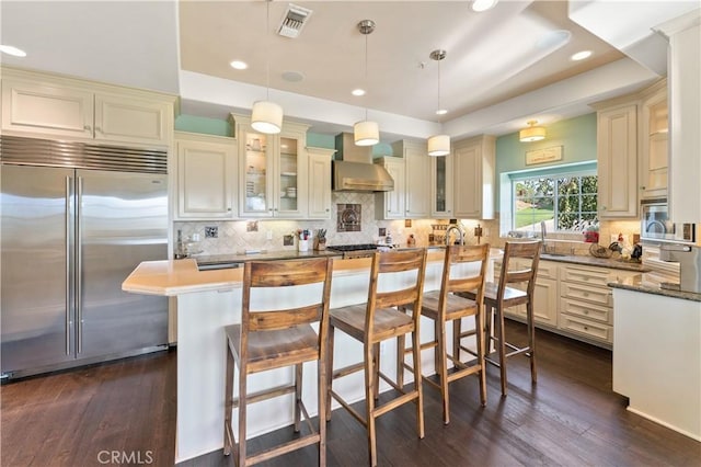 kitchen with pendant lighting, cream cabinets, built in fridge, wall chimney range hood, and a kitchen island