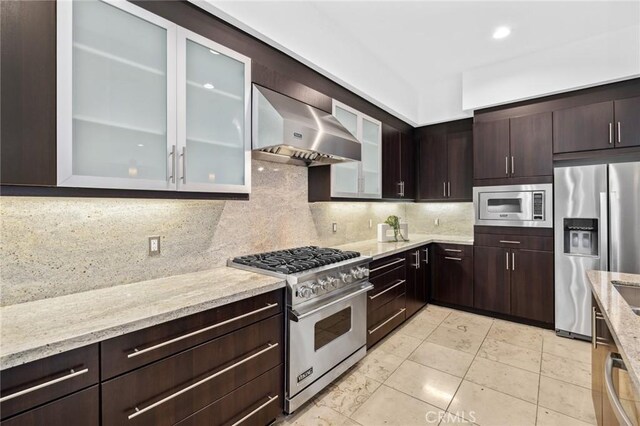 kitchen with backsplash, light stone counters, wall chimney exhaust hood, dark brown cabinetry, and stainless steel appliances