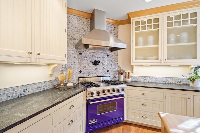 kitchen with wall chimney range hood, dark stone counters, decorative backsplash, premium stove, and cream cabinetry