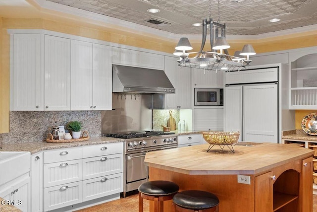 kitchen featuring decorative light fixtures, wall chimney range hood, a center island, built in appliances, and white cabinetry