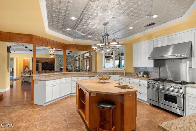 kitchen with pendant lighting, wall chimney exhaust hood, white cabinetry, an island with sink, and double oven range