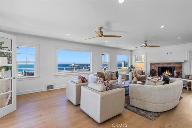 living room with a water view, crown molding, a brick fireplace, ceiling fan, and light wood-type flooring