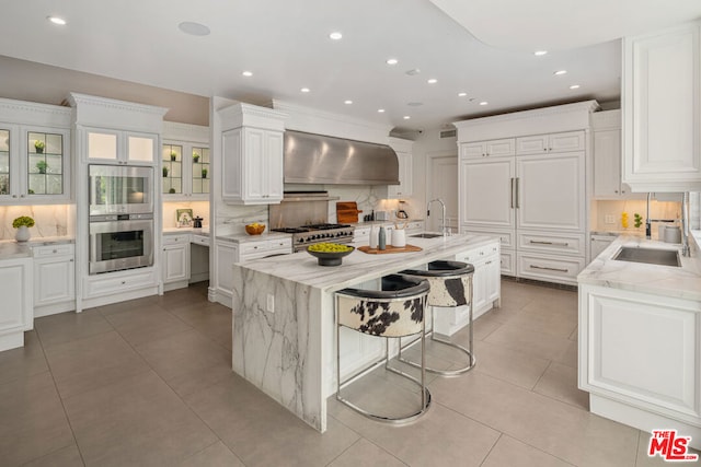 kitchen featuring white cabinetry, light stone countertops, wall chimney range hood, and a kitchen island with sink