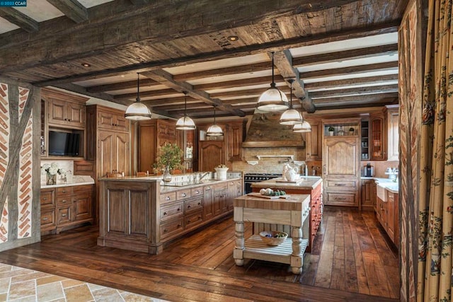 kitchen with premium range hood, decorative light fixtures, beamed ceiling, backsplash, and a kitchen island with sink