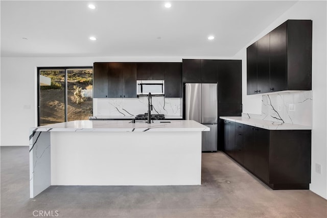 kitchen with light stone countertops, stainless steel refrigerator, a kitchen island with sink, and tasteful backsplash