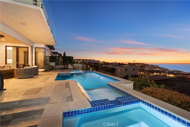 pool at dusk with an in ground hot tub, a water view, and a patio area