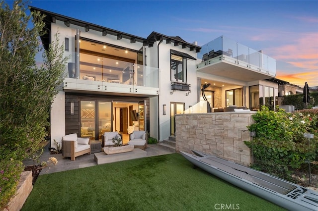 back house at dusk featuring a lawn, a patio area, a balcony, and an outdoor hangout area