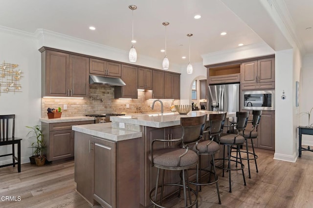 kitchen featuring light stone counters, decorative light fixtures, a center island with sink, stainless steel appliances, and light wood-type flooring