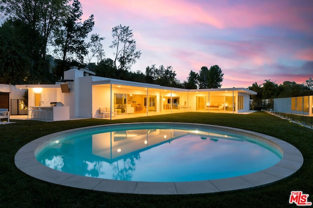 pool at dusk featuring a patio, exterior kitchen, and a lawn