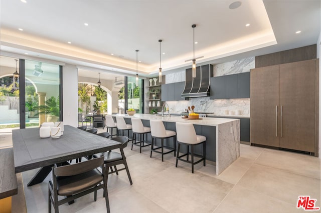 kitchen with wall chimney range hood, an island with sink, decorative light fixtures, a raised ceiling, and tasteful backsplash