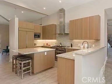 kitchen with wall chimney exhaust hood, built in appliances, a center island, a breakfast bar area, and light wood-type flooring