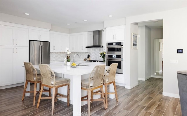 kitchen featuring wood finish floors, stainless steel appliances, backsplash, a sink, and wall chimney exhaust hood