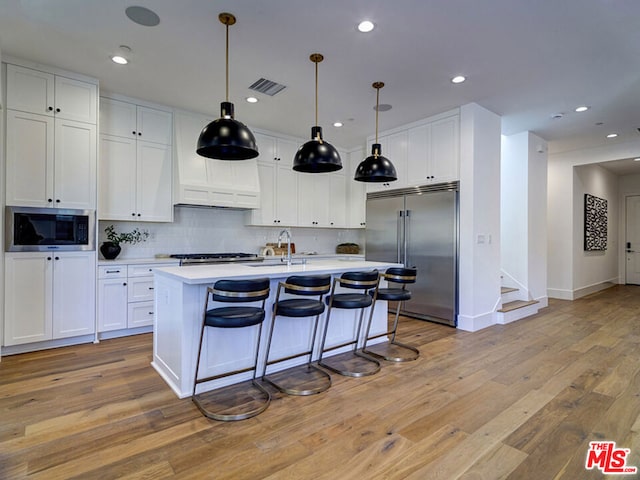 kitchen with white cabinets, an island with sink, built in appliances, and light wood-type flooring