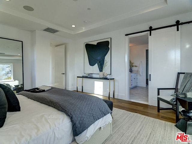 bedroom featuring a barn door, a tray ceiling, ensuite bath, and light hardwood / wood-style flooring