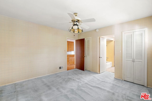 unfurnished bedroom featuring ceiling fan, light colored carpet, ensuite bathroom, and multiple closets