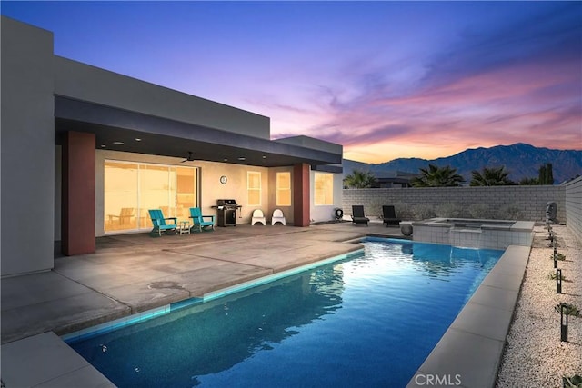 pool at dusk featuring an in ground hot tub, ceiling fan, a patio, a mountain view, and area for grilling