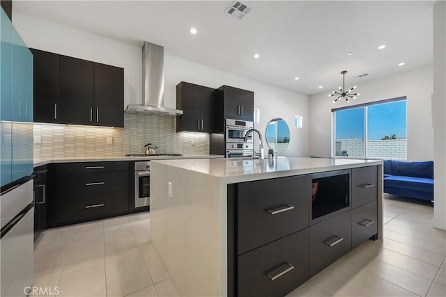 kitchen with pendant lighting, appliances with stainless steel finishes, wall chimney range hood, decorative backsplash, and a kitchen island with sink