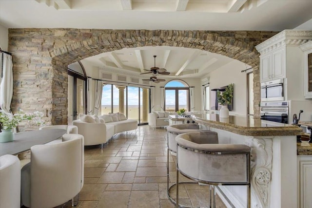 kitchen with stainless steel oven, a breakfast bar area, beamed ceiling, white cabinetry, and ceiling fan