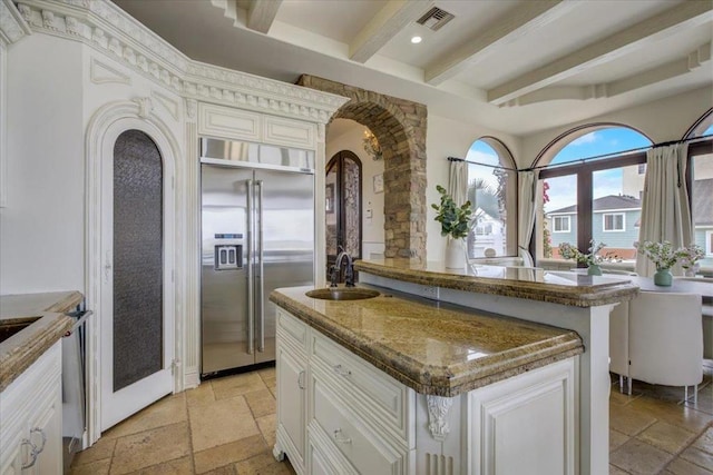 kitchen featuring stainless steel built in fridge, a kitchen island with sink, beam ceiling, dark stone counters, and sink