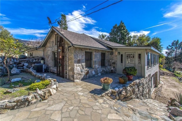 view of front of house with cooling unit, stone siding, a patio area, and roof with shingles