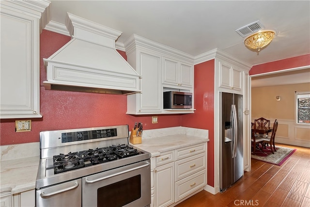 kitchen with appliances with stainless steel finishes, custom range hood, dark wood-type flooring, light stone counters, and crown molding