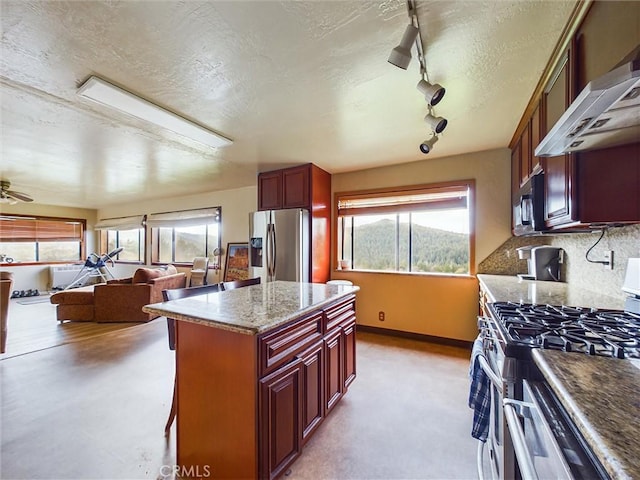 kitchen featuring wall chimney range hood, rail lighting, ceiling fan, dark stone countertops, and appliances with stainless steel finishes