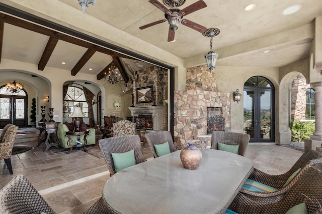 dining space with french doors, lofted ceiling with beams, ceiling fan with notable chandelier, and an outdoor stone fireplace